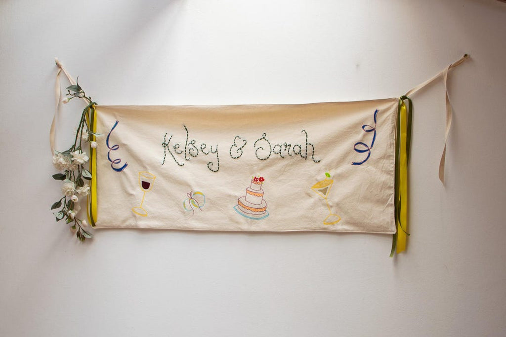 Cream illustrated wedding banner with green embroidered names, painted cake and wine glasses, yellow and green ribbons