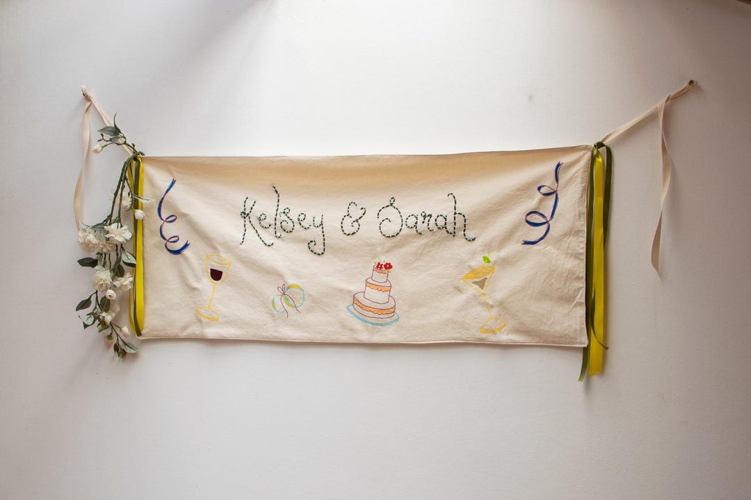 Cream illustrated wedding banner with green embroidered names, painted cake and wine glasses, yellow and green ribbons