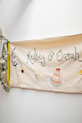 Illustrated wedding banner in natural cotton with hand-embroidered names and hand-painted cake, wine glass and gift