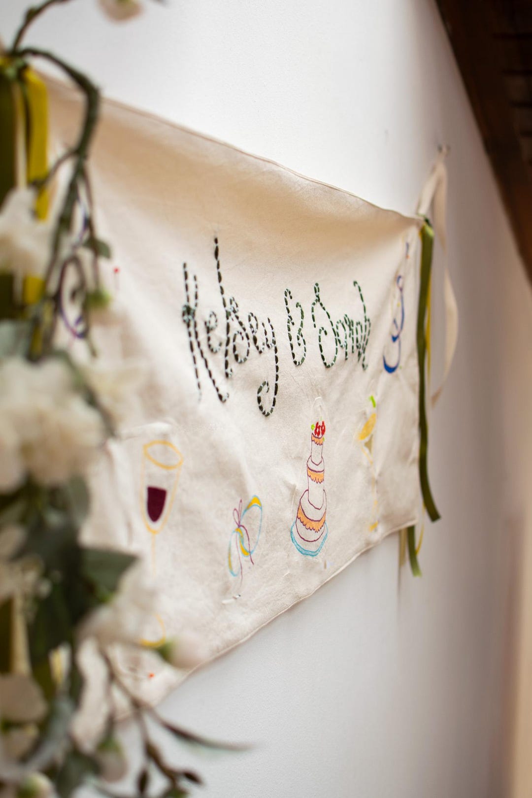 Close-up of cream cotton banner with names embroidered in black thread and hand-painted champagne glass, cake and bow